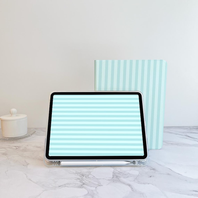 May include: A digital tablet with a light blue and white striped screen, a white stylus, and a light blue striped notebook. The objects are on a marble surface, with a white container with a lid in the background.