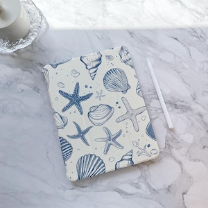 Può includere: Una custodia per iPad bianca con un motivo a conchiglie e stelle marine blu e bianche.