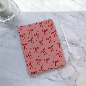 Könnte beinhalten: Eine rosa iPad-Hülle mit einem roten Schleifenmuster. Die Hülle hat ein dezentes Punkte-Muster.