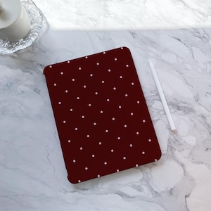 Peut inclure: Une coque de tablette bordeaux ornée d'un motif à pois de petits points blancs espacés. Un stylet blanc est posé sur une surface en marbre à côté de la coque. La coque est rectangulaire avec des coins arrondis.