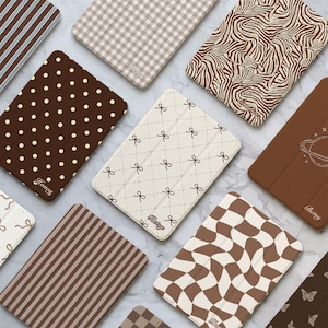 Puede incluir: Una colección de fundas rectangulares para tabletas con varios patrones, incluyendo lunares marrones y blancos, cuadros vichy beige, rayas de cebra, crema con lazos marrones, un gráfico de planeta, rayas marrones y un tablero de ajedrez ondulado.