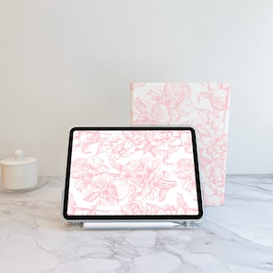 May include: A white tablet with a pink floral design on the screen. The tablet is sitting on a white marble surface. A white notebook with a pink floral design is behind the tablet.