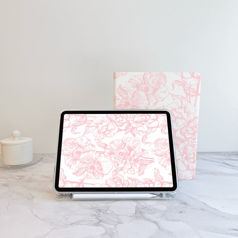 May include: A white tablet with a pink floral design on the screen. The tablet is sitting on a white marble surface. A white notebook with a pink floral design is behind the tablet.