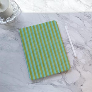 May include: A rectangular tablet case with a green and blue striped pattern. A white stylus is next to the case. The case is on a marble surface.