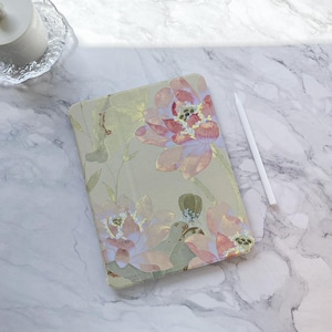 May include: A floral-patterned tablet case featuring large pink and purple flowers on a light green background. A white stylus rests beside the case on a marble surface. The case has a soft, watercolor-like design.
