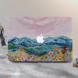 May include: A laptop skin with a painted design of a mountain range with a pink sky and a white apple logo. The mountains are painted in shades of blue, green, and brown. There are small flowers and trees in the foreground.