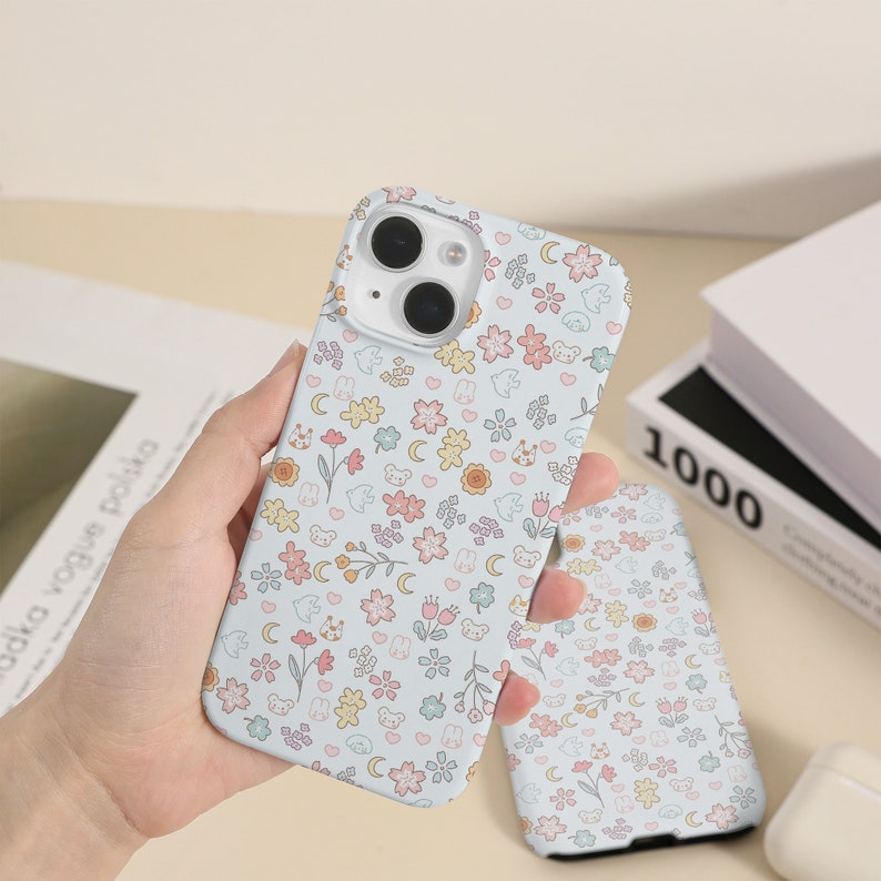 May include: A light blue iPhone case with a cute pattern of flowers, stars, and bears. The case has a clear camera lens.