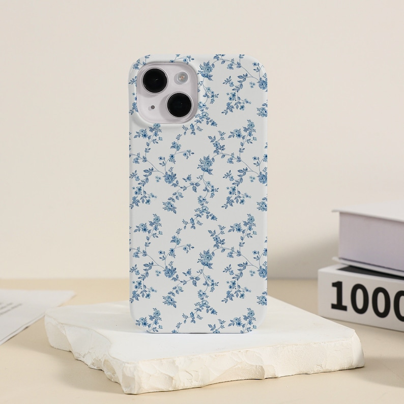 May include: White iPhone case with a blue floral pattern. The case has a cutout for the camera lens.