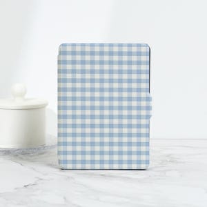 May include: A tablet case featuring a blue and white gingham pattern. The case has a rectangular shape and a magnetic closure. The design is a light blue and white checkered pattern. It rests on a white marble surface.