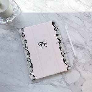 May include: A white iPad case with a black lace design and a black bow on the front. The case is on a white marble surface.