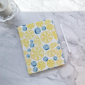 May include: A white tablet case featuring a watercolor pattern of yellow lemon slices and blue blueberries. A white stylus rests on a marble surface next to the case. The case is rectangular and likely designed to protect a tablet.