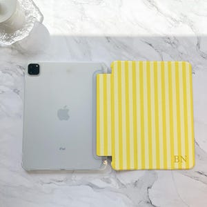 May include: A clear iPad with a yellow and white striped case. The case has the initials "BN" in the bottom right corner. The iPad is on a white marble surface.