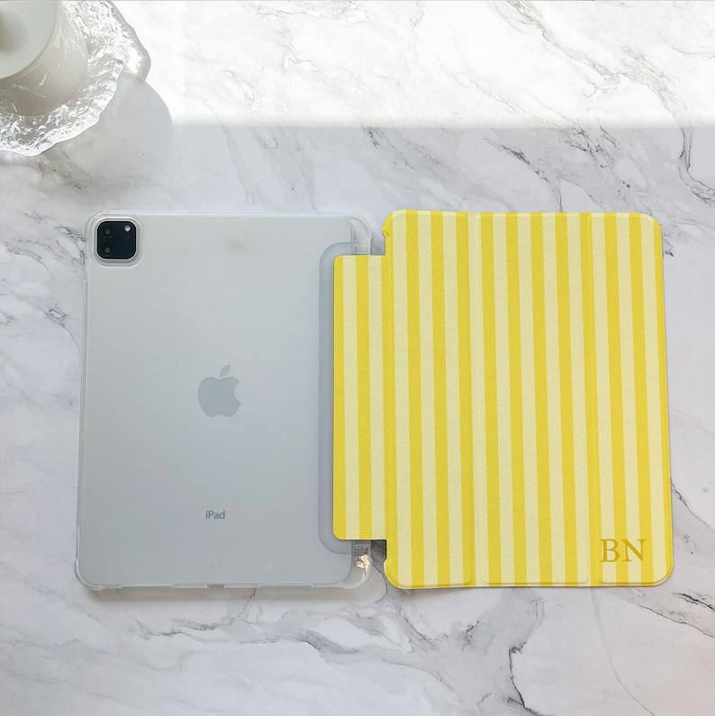 May include: A clear iPad with a yellow and white striped case. The case has the initials "BN" in the bottom right corner. The iPad is on a white marble surface.