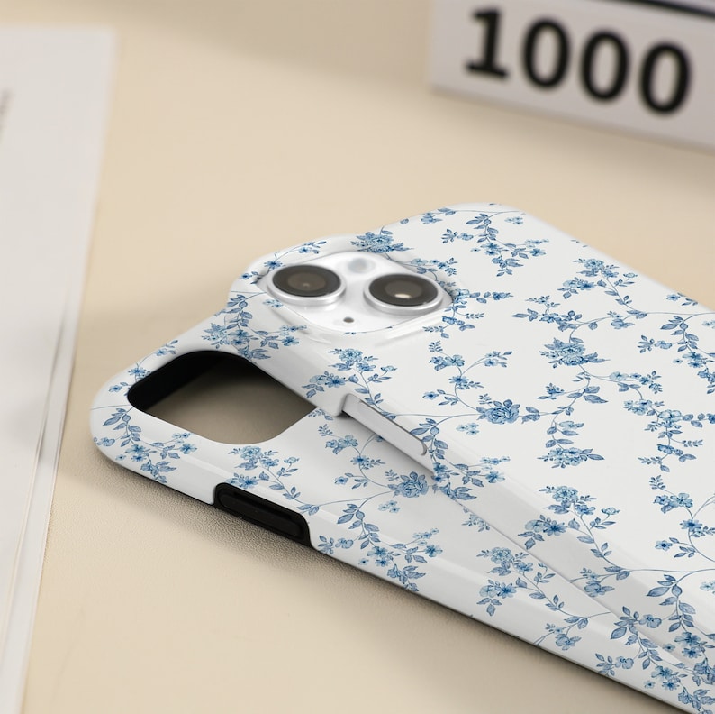 May include: White phone case with a blue floral pattern. The case has a cutout for the camera lens.