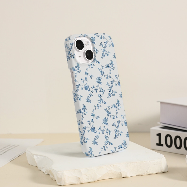 May include: A white phone case with a blue floral pattern. The case has a camera cutout and is designed for an iPhone.