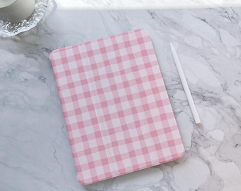 Può includere: Una custodia per tablet a fantasia gingham rosa e bianca con uno stilo bianco. La custodia è rettangolare e si adatta a un tablet. Lo sfondo è una superficie di marmo.