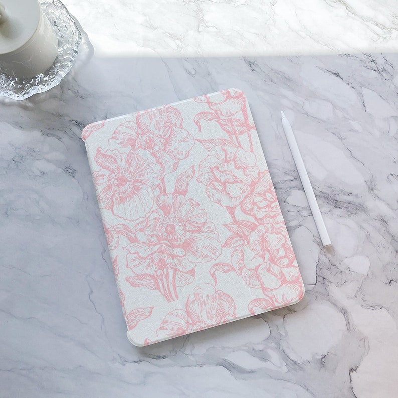 May include: A white iPad case with a pink floral pattern. The case is lying on a white marble surface with a white stylus next to it.