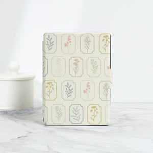 以下が含まれることがあります： クリーム色の電子書籍リーダーカバーに花柄が施されています。パターンは、ピンク、イエロー、グレーの色合いで、繊細な手描きの花と葉が特徴です。カバーは、微妙なエンボス加工が施されています。