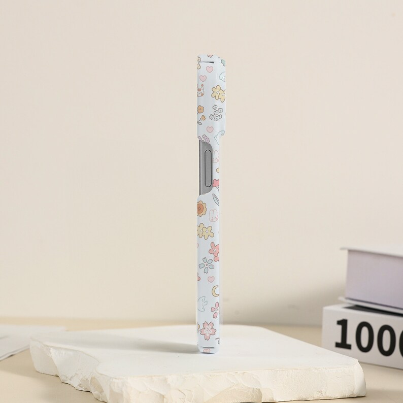 May include: A white phone case with a floral pattern in pastel colors. The pattern includes flowers, hearts, stars, and moons.
