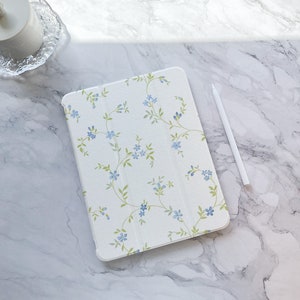 May include: A white iPad case with a floral pattern featuring blue flowers and green leaves. The case is on a white marble surface with a white stylus next to it.