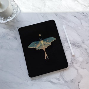 Könnte beinhalten: Schwarze Tablet-Hülle mit einer Illustration einer Mondmotte mit teal- und gelbfarbenen Flügeln und einem kleinen goldenen Stern darüber. Ein weißer Stift liegt auf einer Marmoroberfläche neben der Hülle. Das Design ist zentriert.