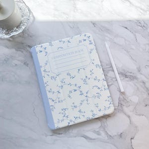Puede incluir: Una funda para iPad de color azul y blanco con estampado floral y el texto "COMPOSITION BOOK" en la parte delantera.