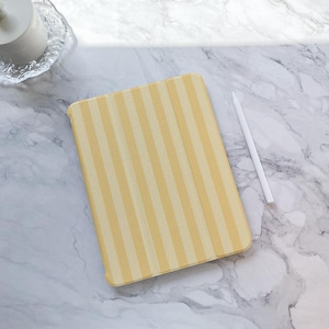 May include: A yellow and white striped tablet case with a matching stylus. The case is rectangular and has vertical stripes. The stylus is white and rests on a marble surface.