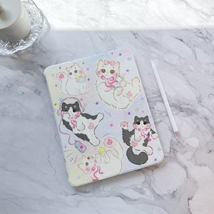 May include: White iPad case with a colorful design featuring multiple cartoon cats wearing headphones, bows, and holding a bottle. The case has a sparkly, iridescent finish.