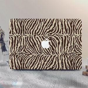 May include: A laptop case featuring a zebra stripe pattern in shades of brown and beige. The Apple logo is visible in the center. The case is on a white surface with other decorative items.