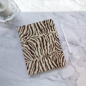 Può includere: Una custodia rettangolare con un motivo zebrato nei toni del marrone e del beige. Uno stilo bianco è posto accanto alla custodia su una superficie di marmo. La custodia è probabilmente per un tablet o un notebook.