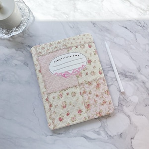 Puede incluir: Un cuaderno de composición blanco con un diseño de patchwork floral rosa. La portada tiene una etiqueta ovalada rosa con el texto "composition book" escrito en negro.