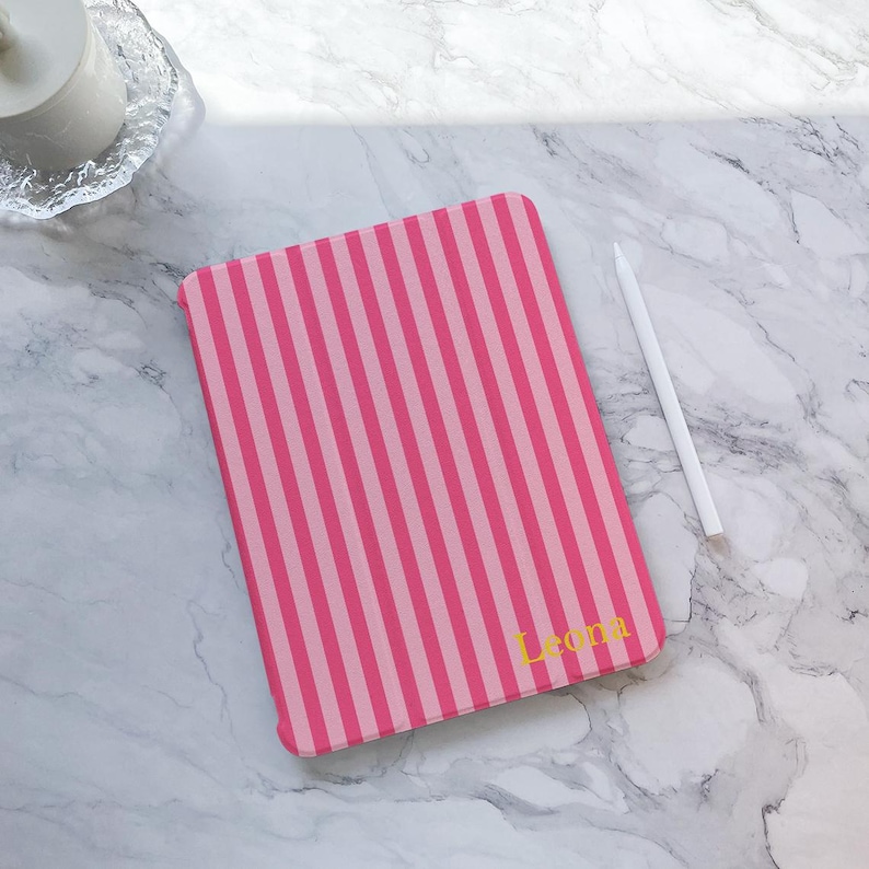 May include: A pink and light pink striped tablet case with the name "Leona" in gold lettering. A white stylus pen rests on a marble surface next to the case. The case is rectangular and has vertical stripes.