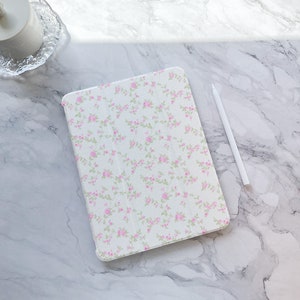 Könnte beinhalten: Eine weiße iPad-Hülle mit einem Blumenmuster aus kleinen rosa Rosen und grünen Blättern. Die Hülle liegt auf einer weißen Marmorfläche mit einem weißen Stift daneben.