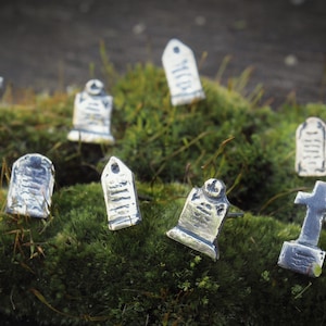 May include: A collection of miniature silver grave markers and crosses, arranged on a bed of green moss. The markers feature various designs, including a cross, a tombstone with an inscription, and a simple rectangular shape.