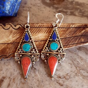 May include: A pair of silver dangle earrings with a triangular design. The earrings feature blue lapis lazuli, turquoise, and coral stones.