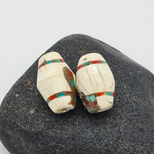 May include: Two white bone beads with turquoise and red inlay designs. The beads are sitting on a dark gray rock.