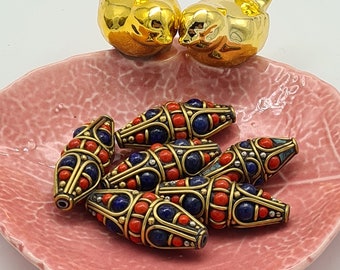 May include: A group of six oval-shaped beads with a brass finish. The beads are decorated with red coral and blue lapis lazuli stones. The beads are arranged in a pink ceramic dish with two gold bird figurines.