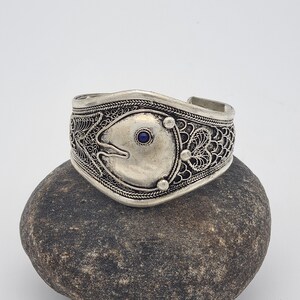 May include: Silver bracelet with intricate filigree design and a blue gemstone accent. The bracelet is resting on a gray rock.