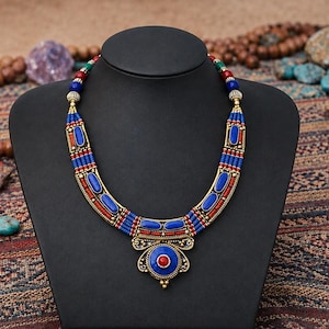May include: A gold-toned metal necklace featuring a central circular pendant with a red stone, surrounded by blue and red cylindrical beads. The necklace has a curved design with alternating blue and red bead segments and gold-toned metal accents.