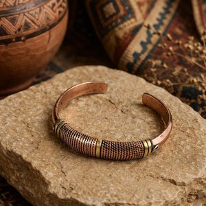 May include: A copper cuff bracelet with a textured central band and gold-coloured accents. The bracelet rests on a light brown stone, with a blurred background featuring a terracotta pot and patterned fabric.
