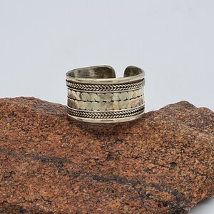 May include: A silver ring with a textured band and a braided design. The ring is sitting on a brown rock.