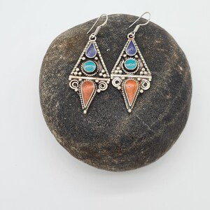 May include: A pair of silver dangle earrings with turquoise, coral, and lapis lazuli stones. The earrings are in the shape of a triangle with intricate details.