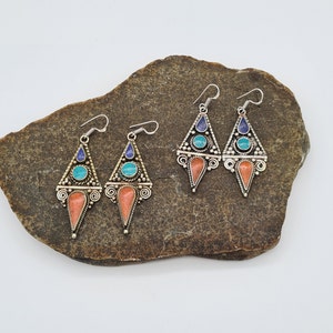 May include: Four silver dangle earrings with turquoise and blue stones and coral accents. The earrings are displayed on a brown rock.