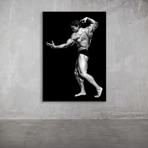 May include: Black and white framed print of a bodybuilder posing against a black background. The bodybuilder is muscular and wearing black briefs. The print is displayed on a gray concrete wall.