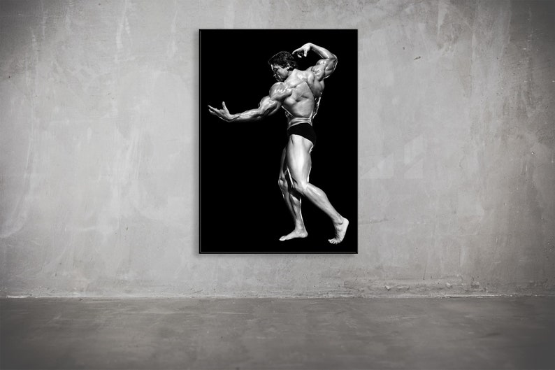 May include: Black and white framed print of a bodybuilder posing against a black background. The bodybuilder is muscular and wearing black briefs. The print is displayed on a gray concrete wall.