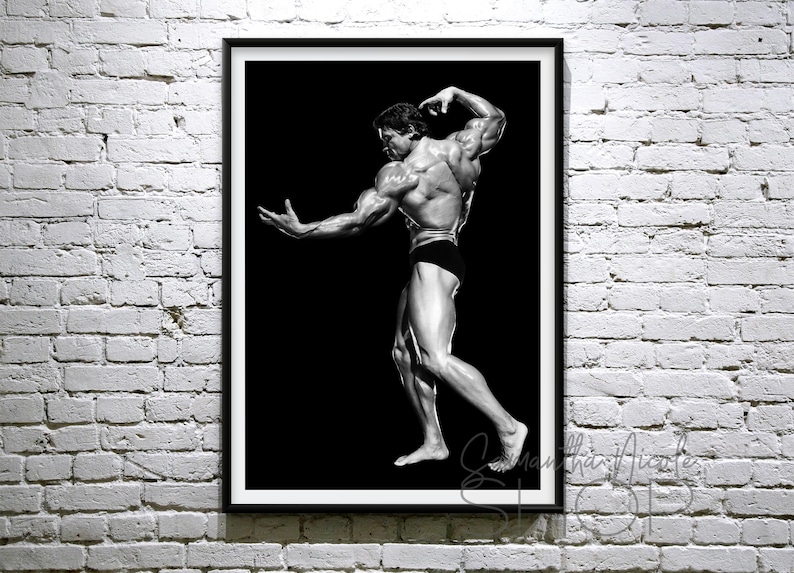 May include: Black and white framed print of a bodybuilder posing. The image features a muscular figure in black shorts against a black background. The print is displayed on a white brick wall.