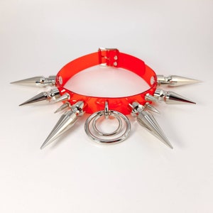 May include: A red plastic choker with silver spikes and a silver ring. The choker is a fashion accessory.