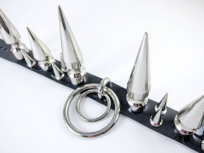 May include: A black leather choker with silver metal spikes and a double ring charm. The choker has a variety of spike sizes.