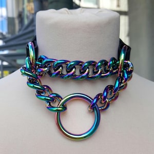 May include: A black choker necklace featuring a chunky, iridescent chain and a large ring pendant. The chain and ring have a vibrant, multi-coloured finish, with blues, greens, and purples. The necklace is displayed on a mannequin.