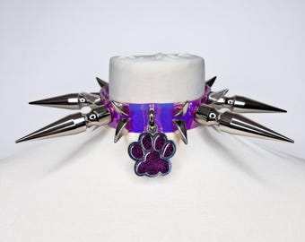 May include: A purple and silver spiked collar with a paw print charm. The collar features large, silver spikes and a shimmering purple band. A small, glittery paw print charm hangs from the center.